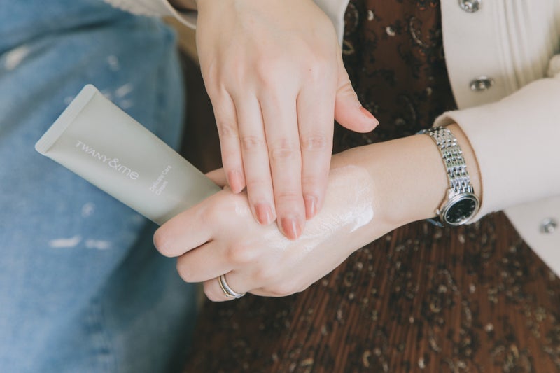 【写真】茶色いワンピースの女性が、チューブタイプの商品を持った左手の甲にクリームを塗っている