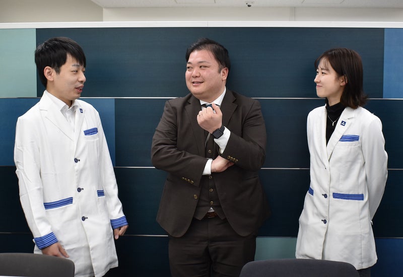 談笑する茶色いスーツの男性と白衣を着た若い男女が3人で談笑している写真
