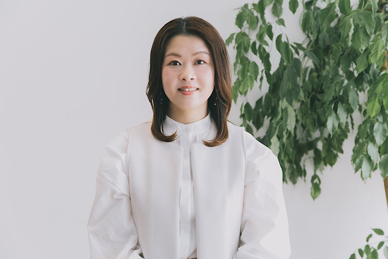 【写真】観葉植物の前に立つ白い服を着た髪の長い女性