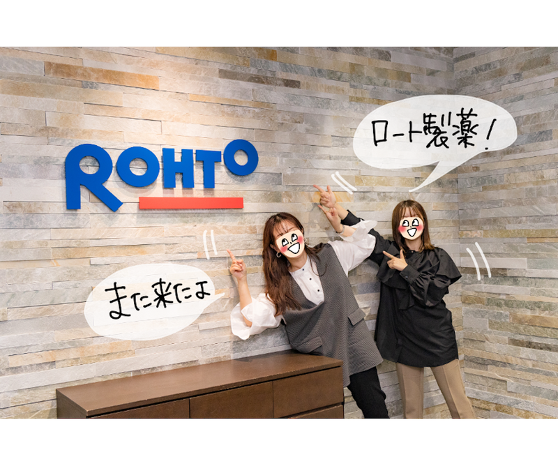 【写真】オギャ子さんとドキ子さんが会社受付でロート製薬のロゴを指さしている